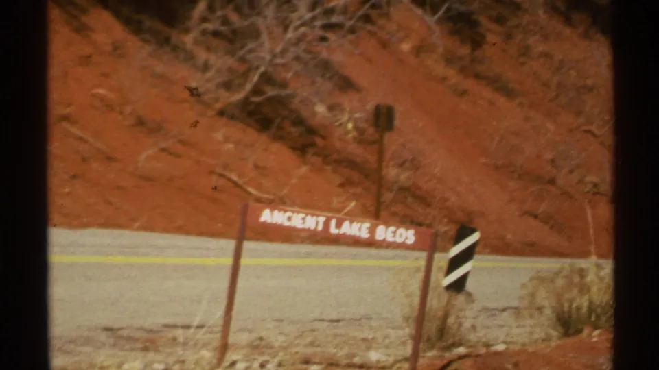 1972 Arizona 8mm frame of the Ancient Lake Beds roadside sign near a paved road, red slope, dry grass, and a striped marker.