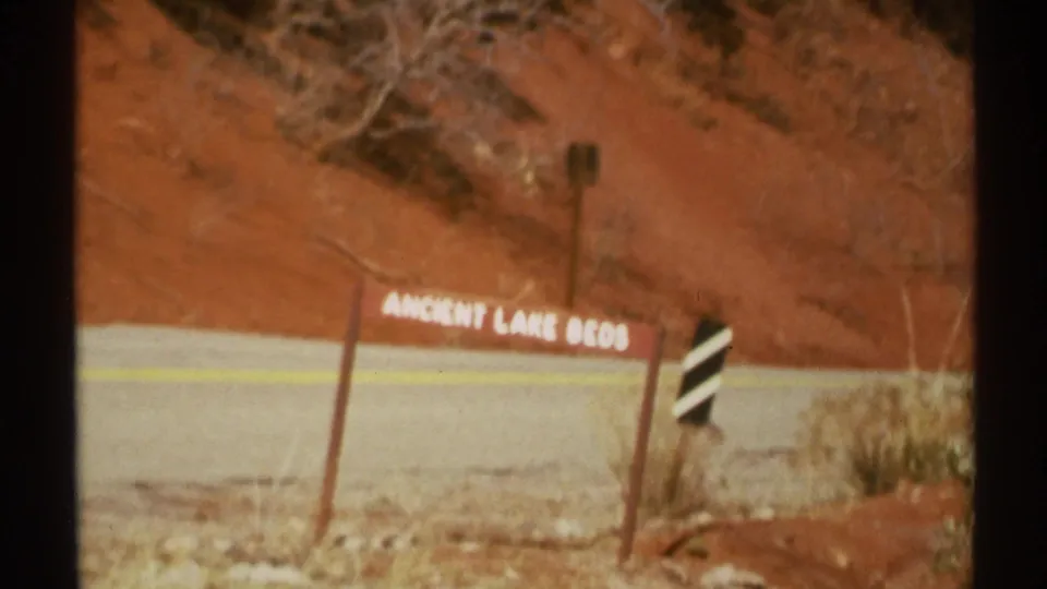 Soft 1972 Arizona 8mm frame showing the Ancient Lake Beds sign, red desert slope, road shoulder, brush, and roadside marker.