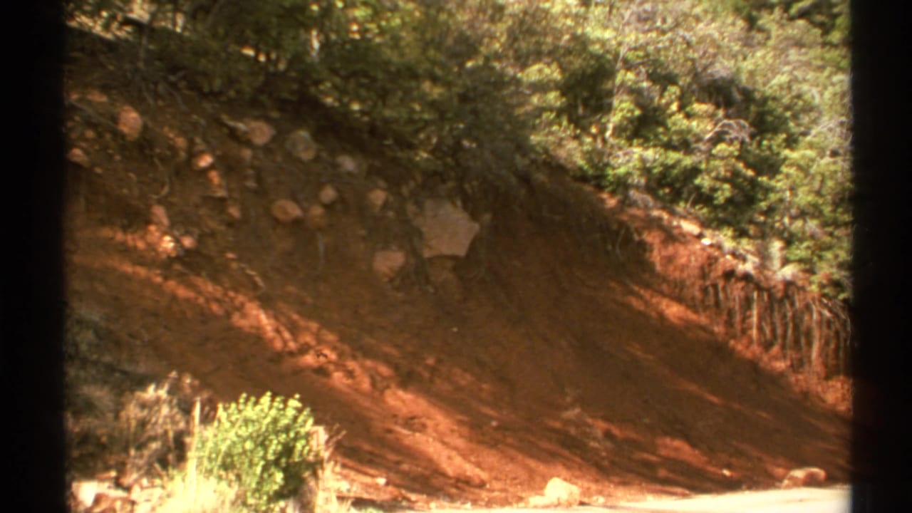 Reddish eroded embankment with shrubs, rocks, and dark film borders.
