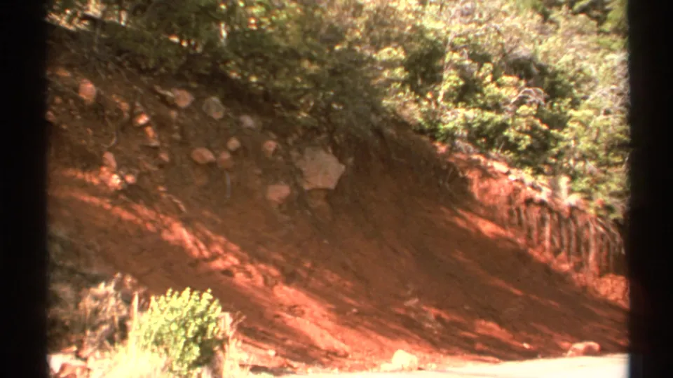 1972 Arizona home-movie frame of a reddish landslide scar with brush above, stones in the soil, and a dark vertical border.