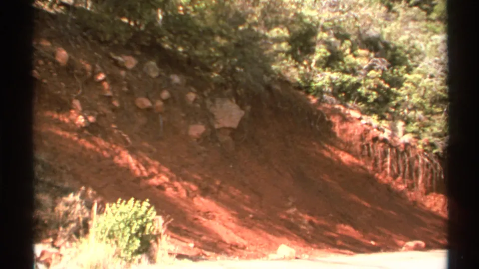 1972 Arizona 8mm frame showing the same eroded red slope, with shrubs at the edge and shadows crossing the exposed soil.