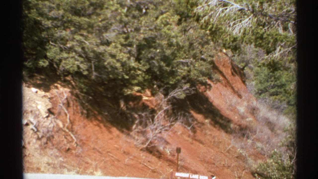 Red eroded hillside with shrubs, dry brush, and a small sign near the lower edge.