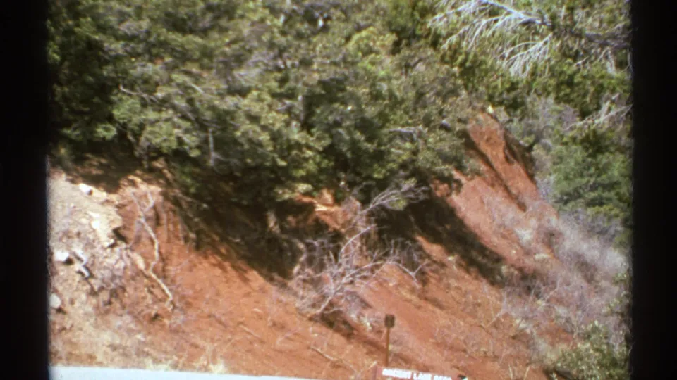 1972 Arizona home-movie frame showing red eroded ground, roadside shrubs, dry brush, and a partly cut-off sign along the lower edge.