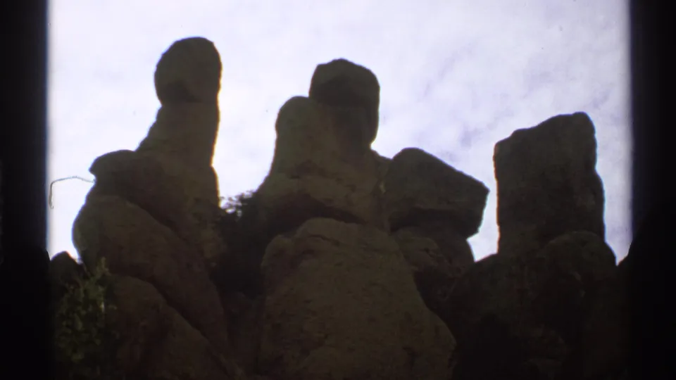 The frame shows several Arizona rock spires and desert shrubs in shadow, filmed on 8mm beneath a pale cloudy sky.