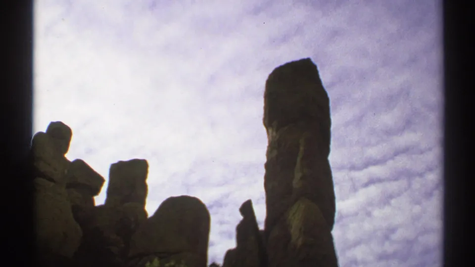 1972 Arizona 8mm frame showing a tall vertical rock formation beside lower boulders and a textured cloudy sky.