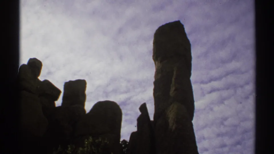 Arizona home-movie frame with irregular stone columns and a taller spire silhouetted against clouds.