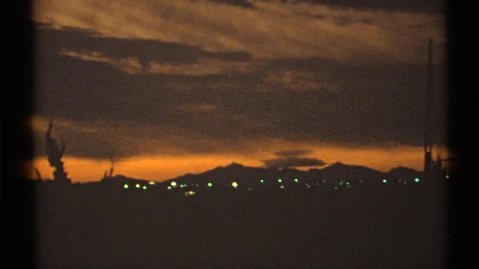 1972 Arizona 8mm frame showing an orange sunset sky, silhouetted mountains, small city lights, and utility poles on the horizon.