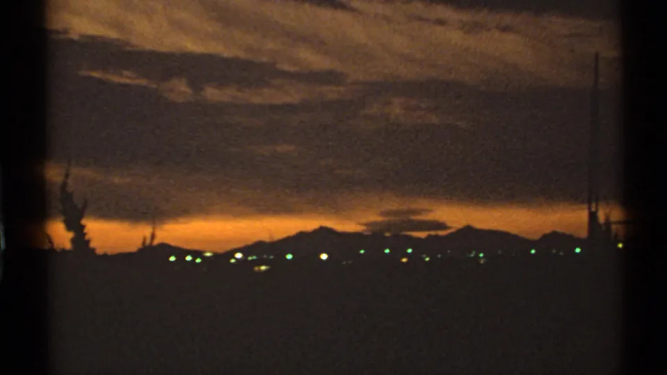 1972 Arizona 8mm frame of a darkening skyline with silhouetted peaks, distant lights, utility poles, and heavy sunset clouds.