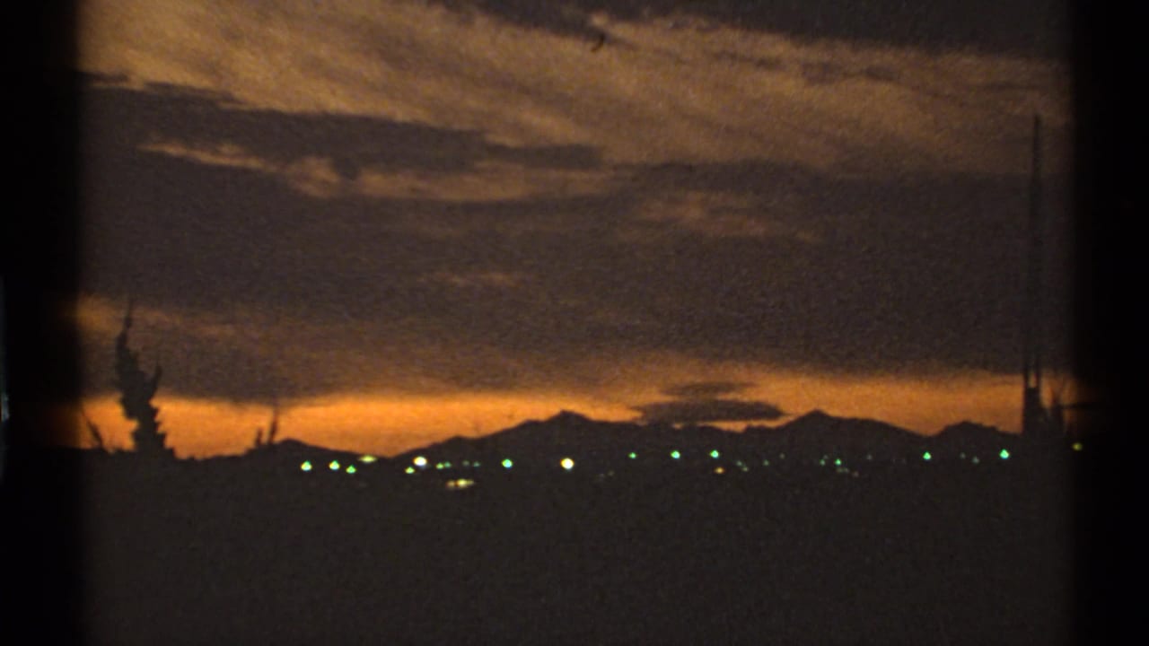 1972 Arizona 8mm frame showing a golden sunset over a distant town, with mountains, clouds, and dark tree silhouettes.