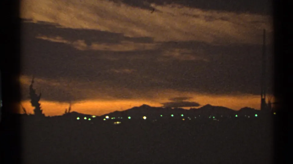 1972 Arizona 8mm frame showing a golden sunset over a distant town, with mountains, clouds, and dark tree silhouettes.