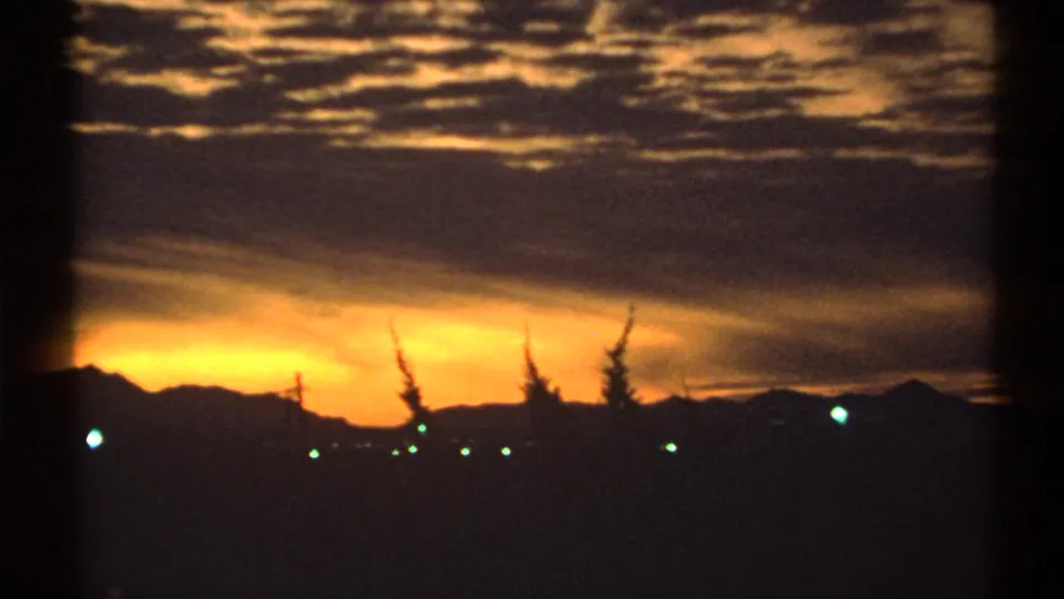 1972 Arizona home-movie frame with orange dusk light, a mountain silhouette, dark trees, and scattered town lights.