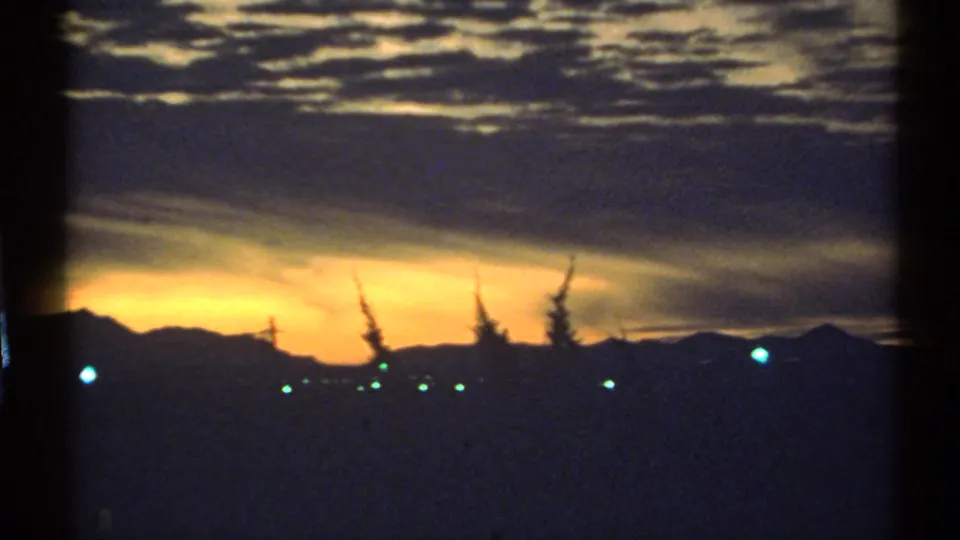 Arizona 1972 8mm sunset frame showing a dark ridge, glowing sky, silhouetted trees, and distant lights at dusk.