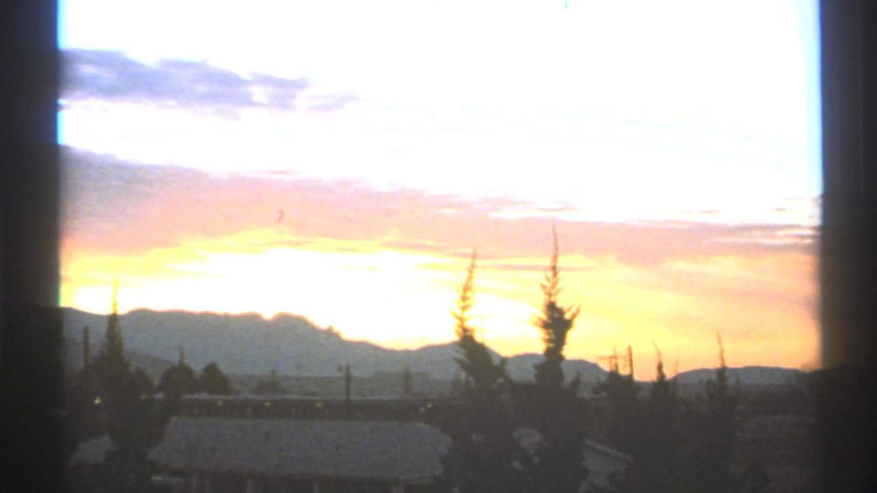 Bright sunset sky over mountains with trees and low structures in silhouette.