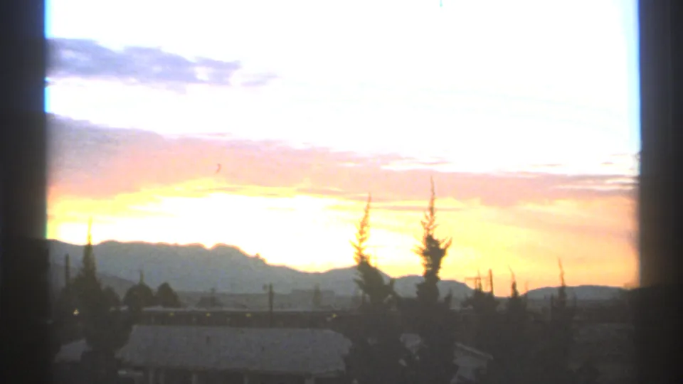 1972 Arizona 8mm frame with a bright orange and pale blue sunset behind a mountain horizon, silhouetted trees, and low distant structures.