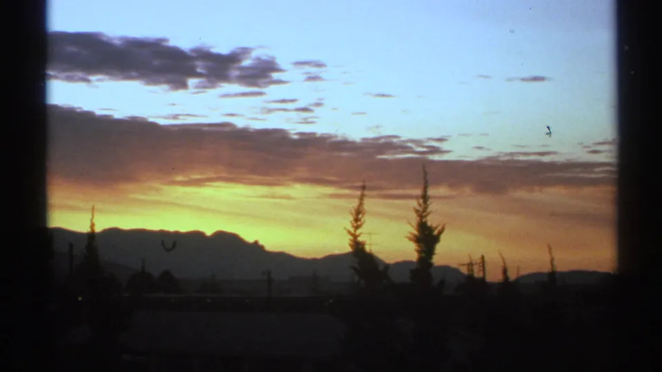 1972 Arizona 8mm frame showing rolling clouds across a blue-orange evening sky, with mountains, trees, and shadowed foreground below.