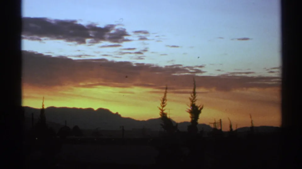 1972 Arizona 8mm frame of a fading sunset over silhouetted mountains, with scattered clouds, tall trees, and a dark foreground.