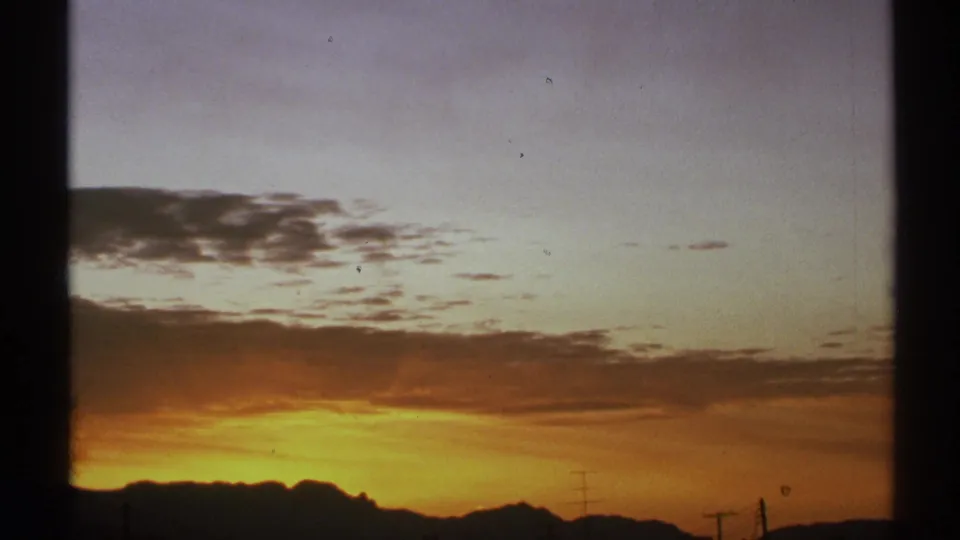 1972 Arizona 8mm frame with orange-yellow dusk light under clouds, dark mountain ridges, and visible utility poles.