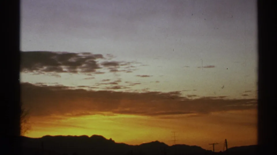 1972 Arizona 8mm frame with fading orange dusk light, cloud texture, mountain silhouettes, and small utility poles.