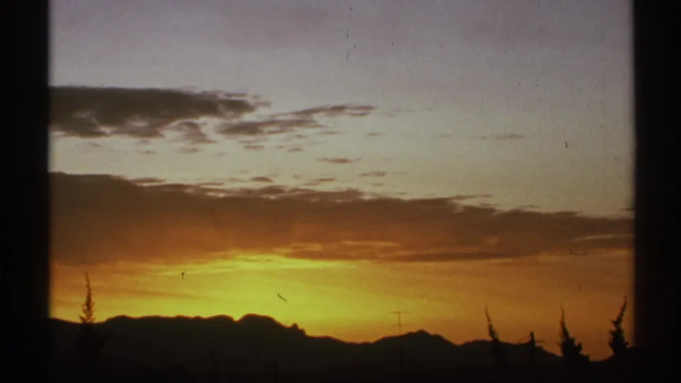 1972 Arizona 8mm frame with a warm sunset sky, silhouetted mountain ridge, small utility poles, trees, and dark window framing.