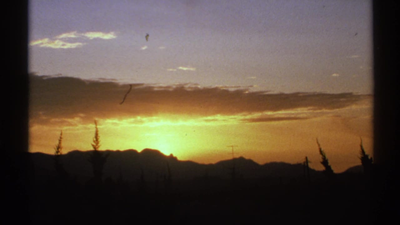 1972 Arizona 8mm frame showing the sun setting behind silhouetted mountains, with orange sky, clouds, and dark foreground trees.