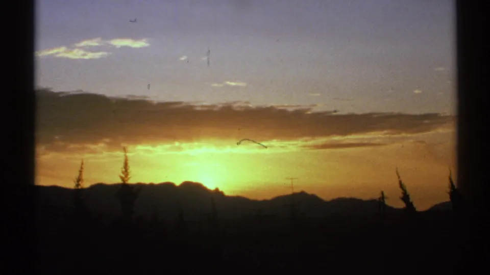 1972 Arizona home-movie view of a bright sunset behind a mountain range, framed by silhouetted trees and a warm clouded sky.