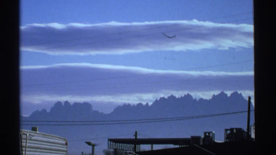 1972 Arizona frame showing layered clouds over a jagged mountain skyline, with power lines and low foreground buildings below.