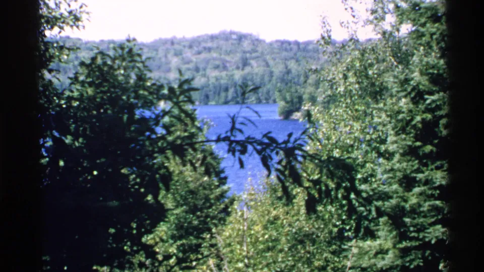 1972 Isle Royale frame showing a blue lake, forested hillside, and shaded evergreen branches in the foreground.