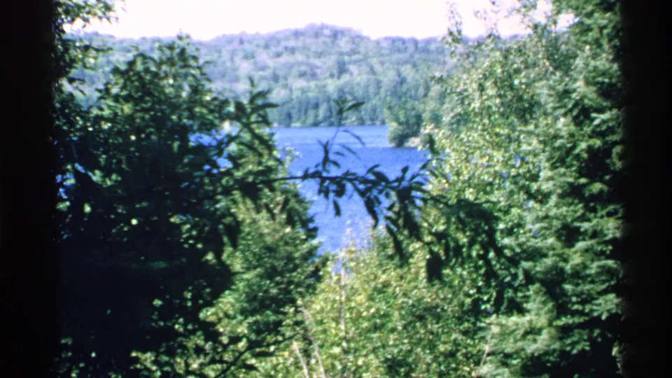 1972 Isle Royale 8mm frame of lake water glimpsed between evergreens and leafy trees, with wooded hills beyond.