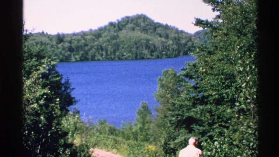 1972 Isle Royale landscape with a blue lake, forested hill, foreground trees, and a light-shirted adult partly visible at the lower right.