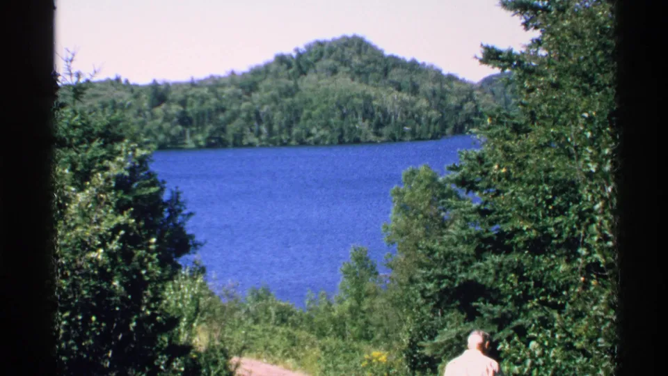Isle Royale home-movie frame showing blue water, dense trees, wooded hills, and an adult visitor seen from behind near the bottom right.