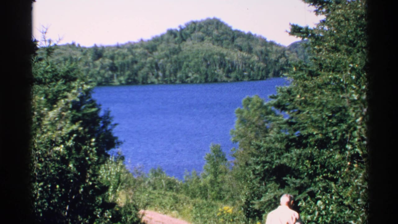 1972 Isle Royale home-movie frame showing a blue lake, wooded hills, dense trees, and a distant adult man near a path.
