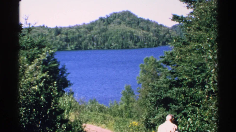 1972 Isle Royale home-movie frame showing a blue lake, wooded hills, dense trees, and a distant adult man near a path.