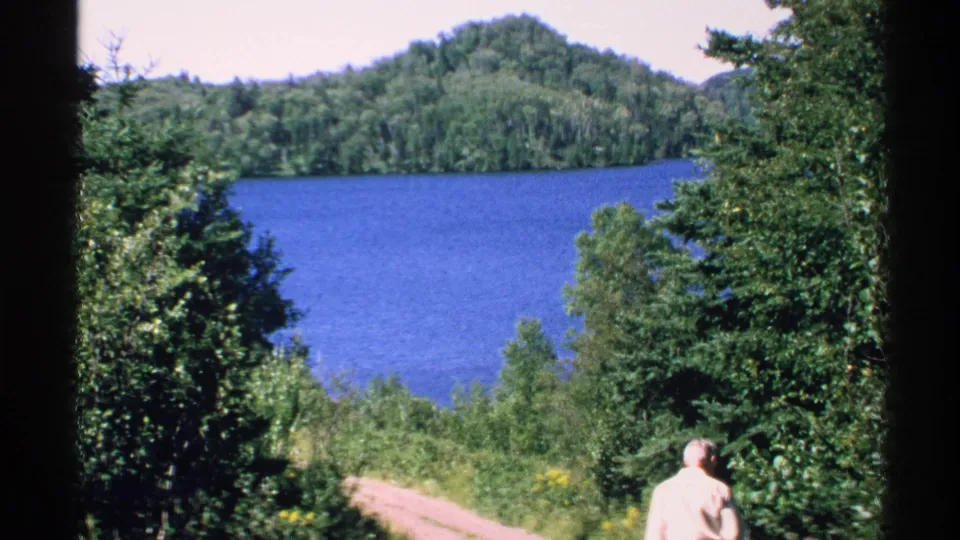 1972 Isle Royale lake view framed by green trees, with a distant adult man in a light shirt near the right edge.