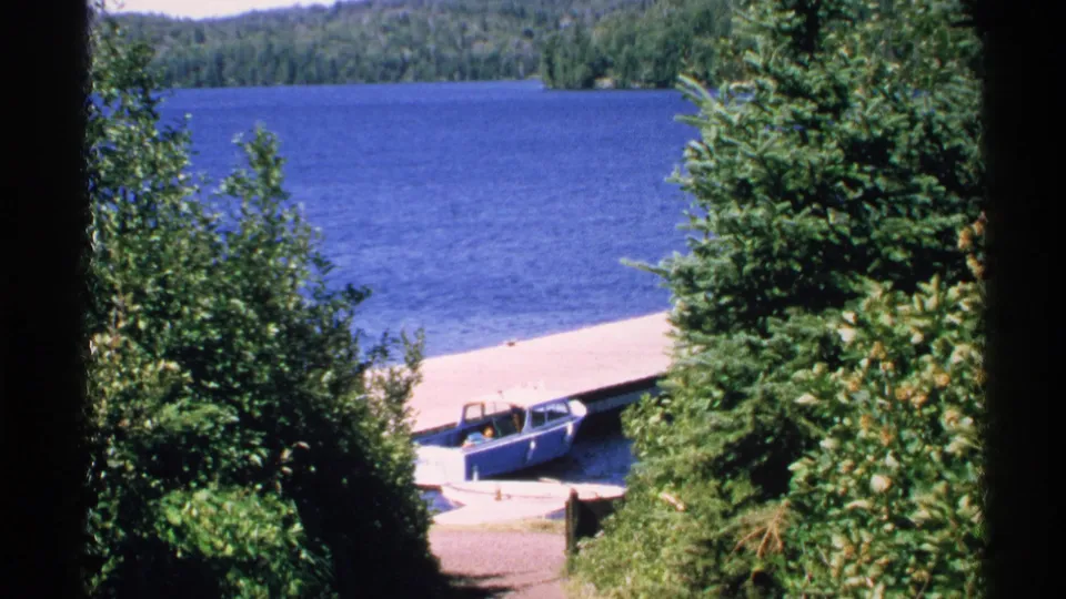 1972 Isle Royale home-movie view of a blue boat beside a dock, framed by dense green trees and lake water.