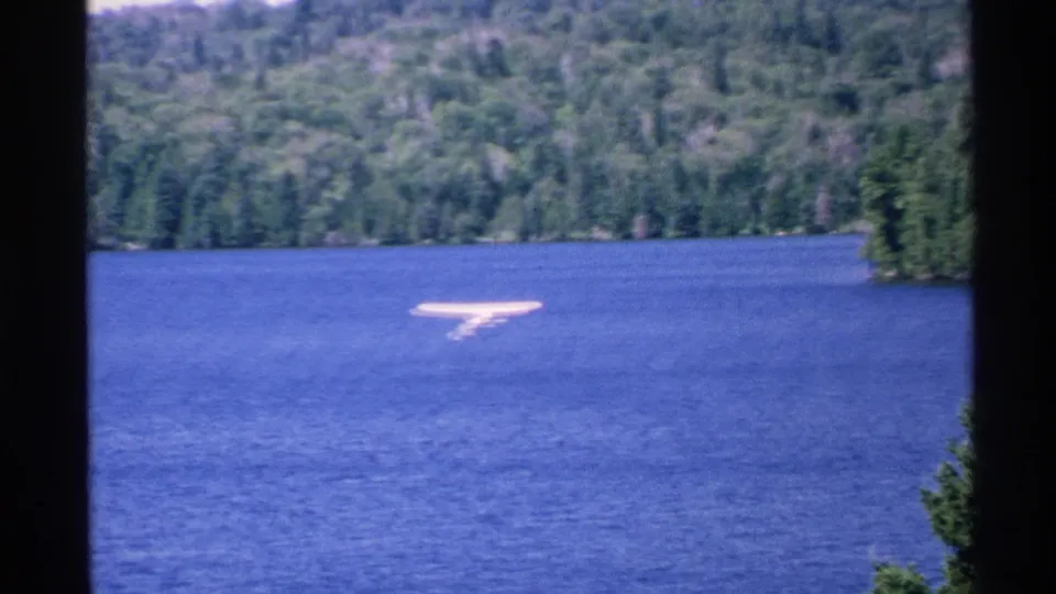1972 Isle Royale home-movie frame showing a small white seaplane low over the lake with forested shoreline and hills in the background.