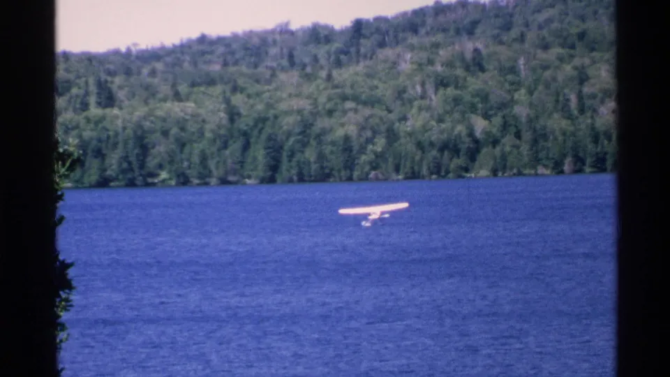 1972 Isle Royale view of a white seaplane moving across open blue lake water, backed by dense green forest and low hills.