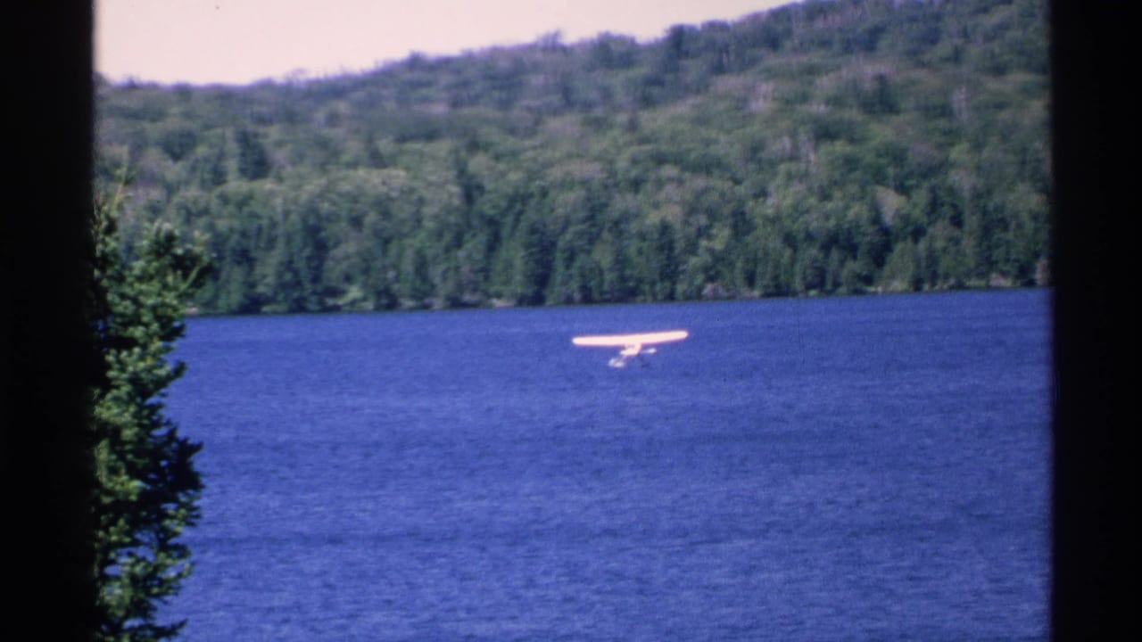 Small floatplane flying low over a blue lake with forested shoreline behind it.