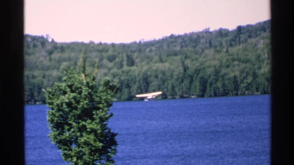 A 1972 Isle Royale frame shows the floatplane nearer the wooded shore, with an evergreen tree in the foreground.