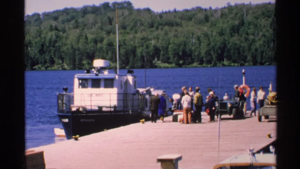 At Isle Royale in 1972, visitors gather on a pier beside a moored boat, vehicles, and a blue forest-lined lake.