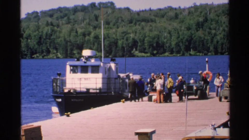 People in 1970s casual clothing stand beside the docked U.S. Mail boat on Isle Royale, with lake water and trees behind them.
