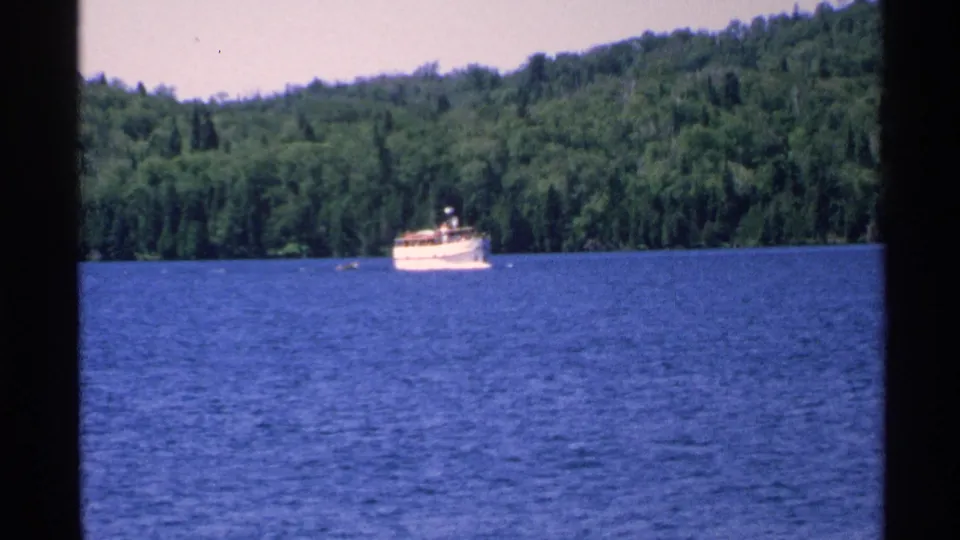 A distant white boat travels across blue water at Isle Royale, framed by the forested shoreline in 8mm home-movie footage.