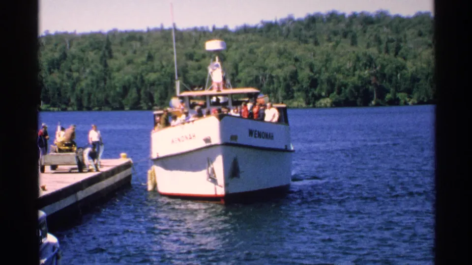The white WENONAH tour boat sits at the dock while people stand along the pier on a wooded Isle Royale lakefront.