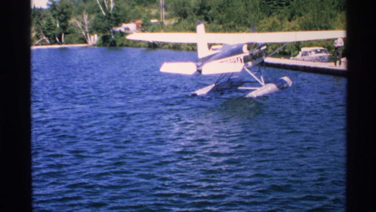 1972 Isle Royale frame with a high-wing pontoon seaplane near a dock, forested shore, parked car, and distant person.