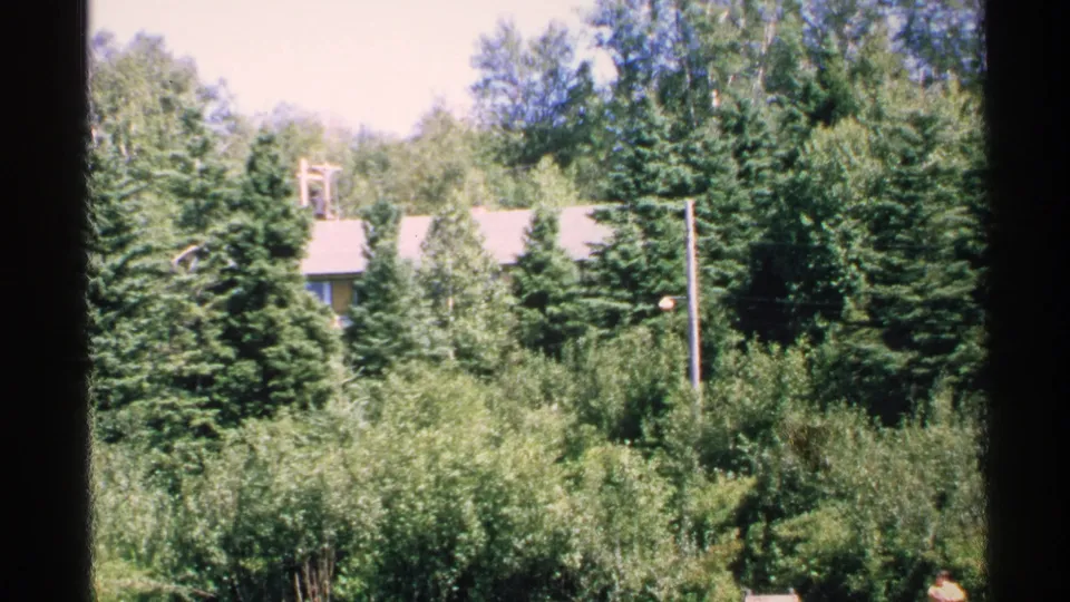 1972 Isle Royale 8mm frame looking toward dense trees and a low cabin roofline beyond brush.