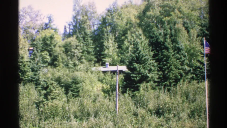 1972 Isle Royale home-movie frame with a dense green tree line, concealed roof, slim pole, and American flag at the right side.
