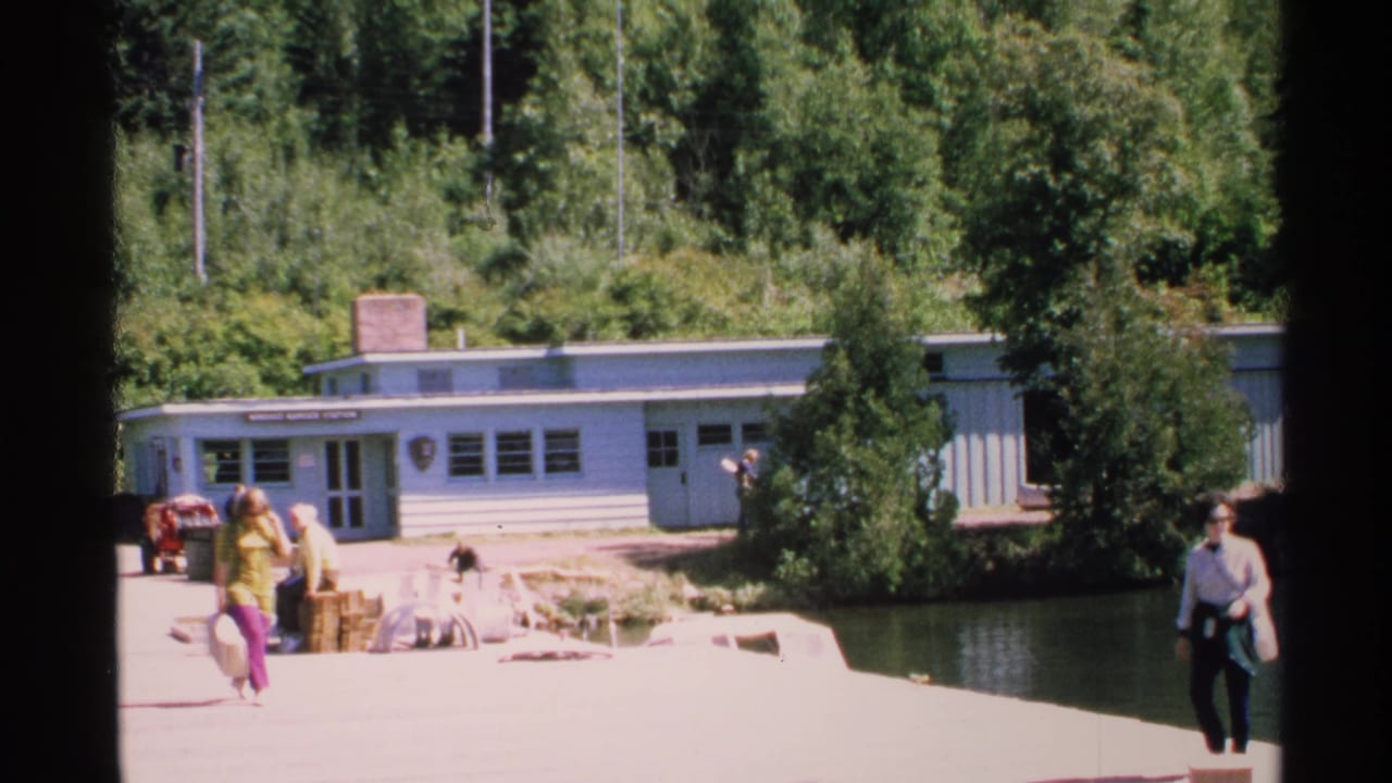 Tourists walk with luggage on a sunny dock near a light blue lakeside visitor center.