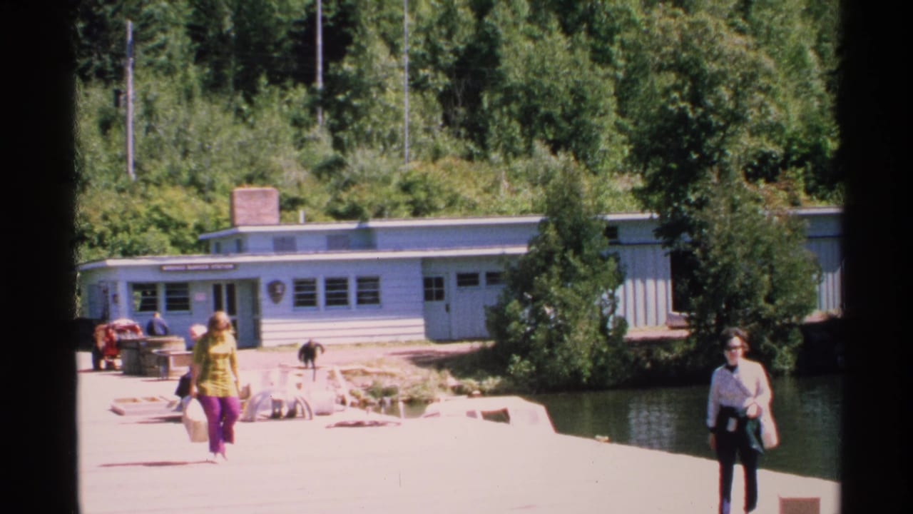Visitors cross a dock beside water and a low visitor center building in bright sun.