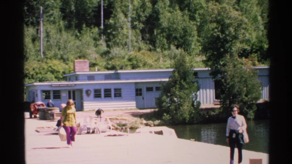 Tourists at Isle Royale's Windigo area walk near a light blue visitor center and lakeside dock in 1972 home-movie footage.