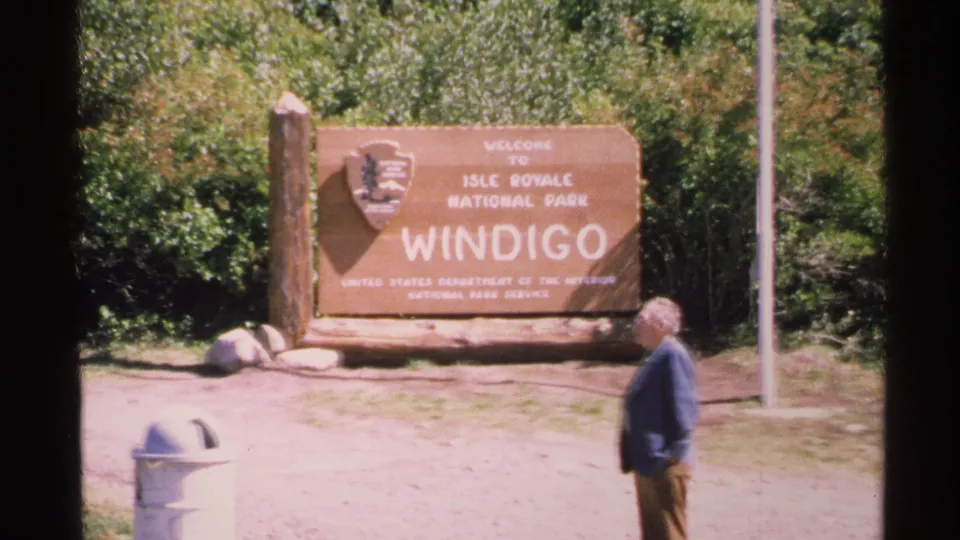 A visitor stands in front of the Welcome to Isle Royale National Park Windigo sign, with trees and a flagpole nearby.
