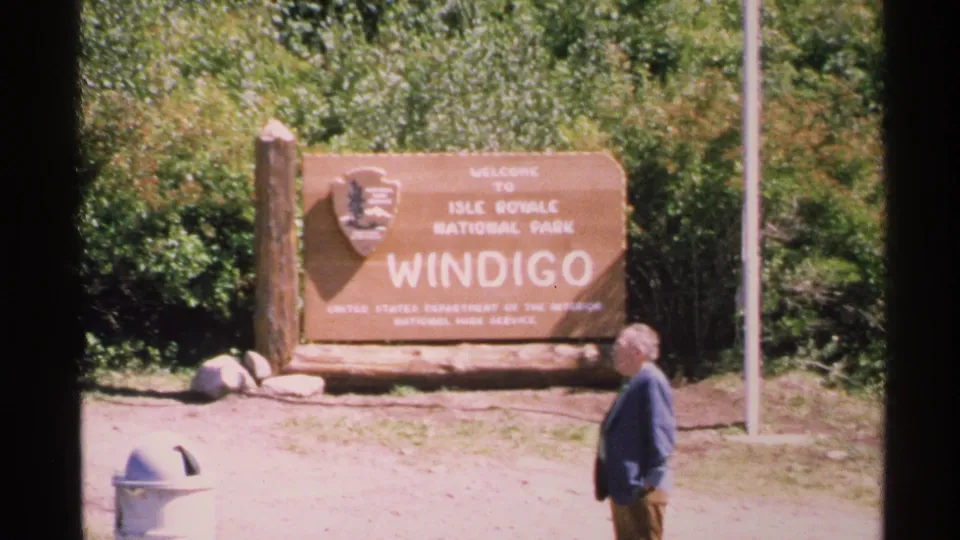 The Windigo sign for Isle Royale National Park fills the frame as a man passes in front of it in 1972 footage.